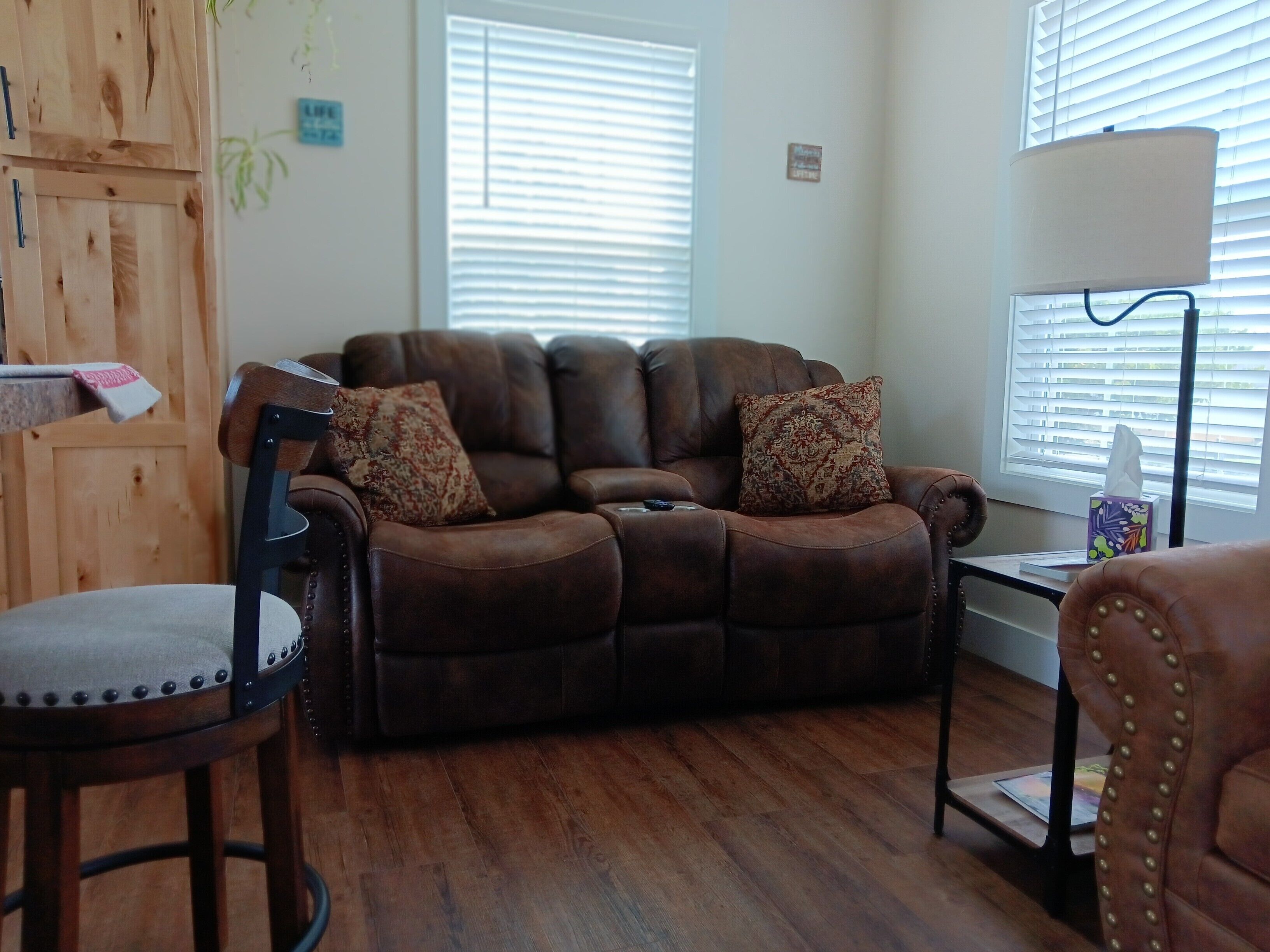 A living room with a brown leather sofa, two patterned throw pillows, a wooden chair, a small side table with a lamp, and a window with blinds.