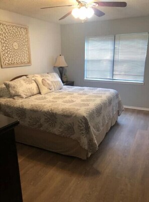Relax in this serene bedroom with a large bed and ceiling fan, featuring a window with blinds and a decorative panel.