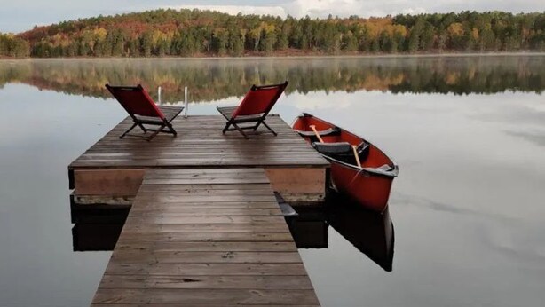 A wooden dock extending into calm waters with two red chairs and a red boat.