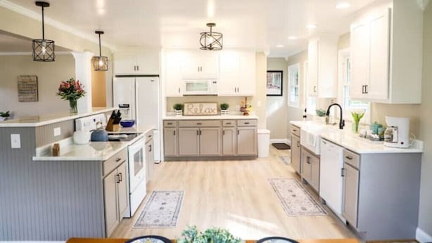 Une cuisine moderne avec des meubles blancs, un îlot central, des luminaires suspendus et un panneau « Détendez-vous » accroché au mur.