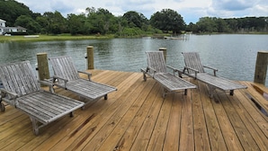 Une terrasse en bois avec quatre chaises en métal, donnant sur un lac, entourée de verdure et avec une maison en arrière-plan.
