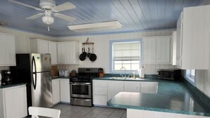 Cocina con alacenas blancas, un refrigerador de acero inoxidable, estufa y microondas. Se ve una ventana con persianas, un ventilador de techo y piso de baldosas.