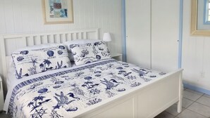 Un dormitorio con una cama blanca, ropa de cama estampada en azul y blanco, un cuadro enmarcado en la pared y una puerta cerrada con detalles azules.