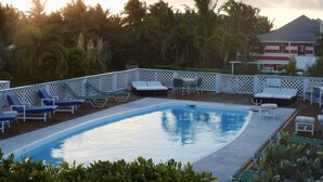 Una piscina con sillas de descanso y una cerca blanca, rodeada de palmeras y un edificio al fondo.