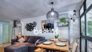 En moderne stue med sofa, spisebord med stole, en hængende lampe og et stort vindue.