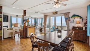 A spacious living area with a wooden dining table, comfortable seating, a ceiling fan, and large windows offering a view of the ocean. - Beautiful Oceanfront Beach House for the whole family and more (Bethany Beach)