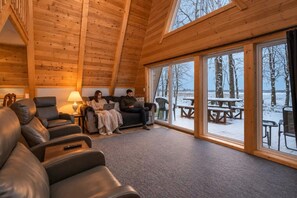 A wooden cabin interior with a sofa, chairs, and a view of a snowy outdoor area with picnic tables.