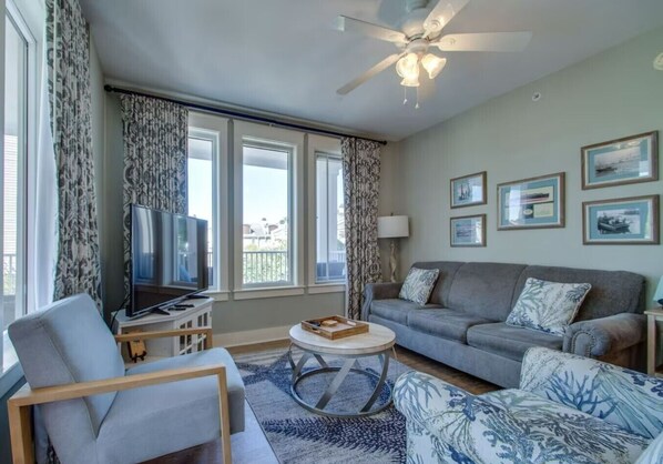 A living room with a grey sofa, a TV, a ceiling fan, and a large window with curtains. - 2 BR/2 BA With 6 Seater Golf Cart, Village of Baytowne Wharf (Miramar Beach)