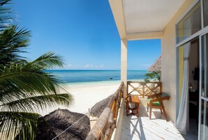 Vista a la playa desde un balcón con techo de paja, una mesa y sillas de madera.
