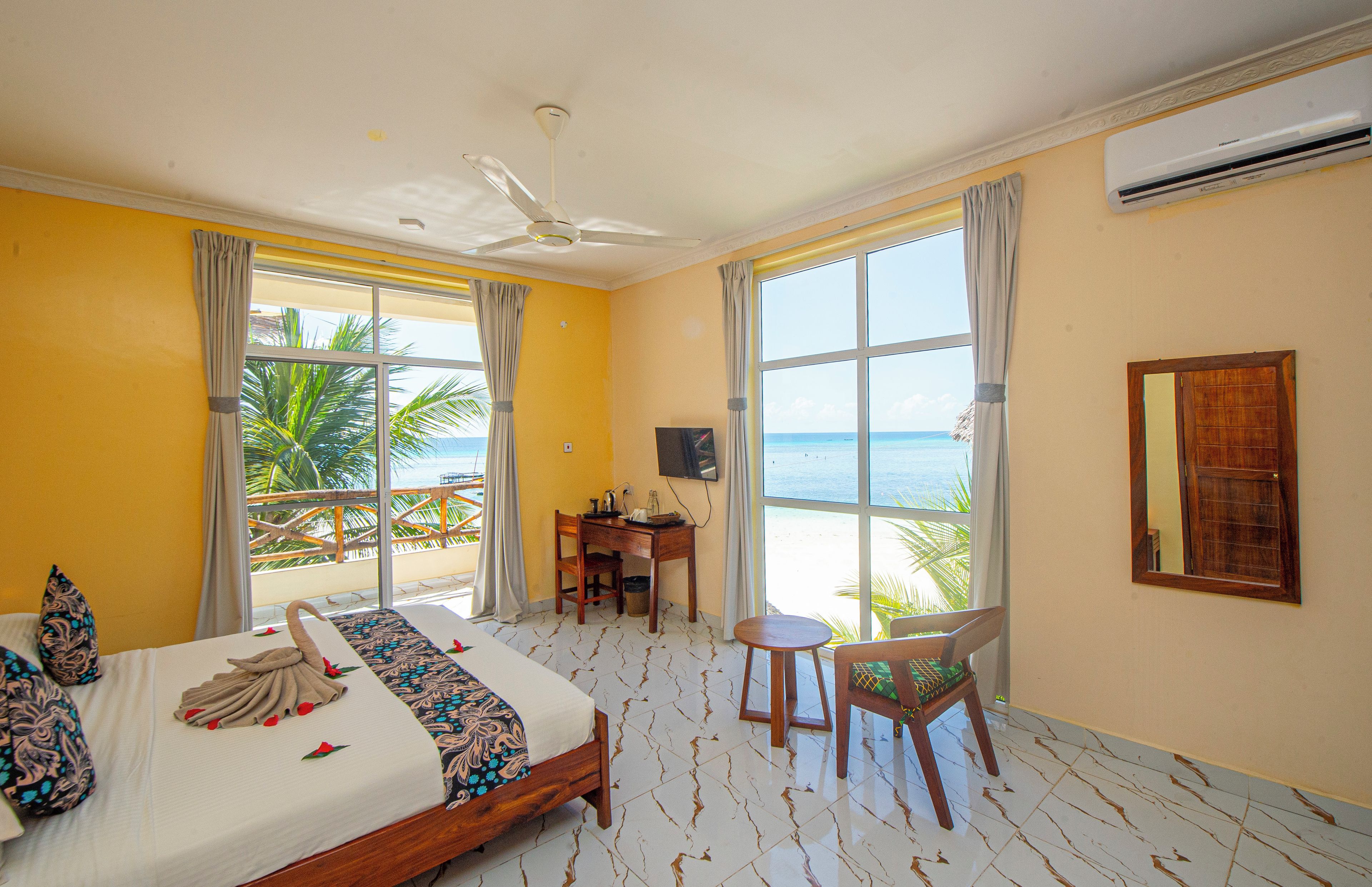 A hotel room with a bed, a desk, a chair, and large windows overlooking the sea.