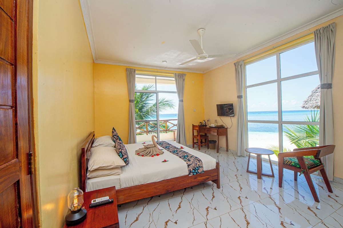 A hotel room with a bed, a desk, a chair, a ceiling fan, and a large window with a view of the beach.