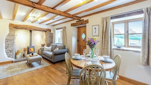 Een gezellige woonkamer met een stenen openhaard, houten balken, een gedekte eettafel voor vier personen en uitzicht door een raam.