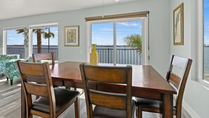 Un comedor con mesa y sillas de madera, ventanas amplias con vista al agua y un ventilador de techo.
