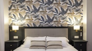 A double bed with white linens and two pillows, flanked by nightstands with lamps, against a wall with a tropical leaf pattern. - Hola Bilbao! (Bilbao)