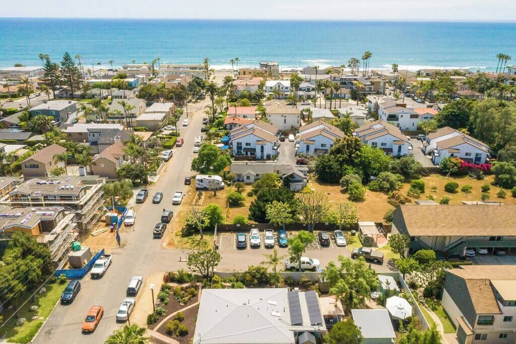 An aerial view of a coastal residential area with houses, a parking lot, and a beach in the background.