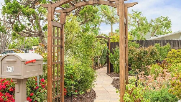 Une tonnelle en bois avec une boîte aux lettres, entourée d’un jardin où poussent différentes plantes et bordée d’un chemin en pierre.