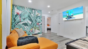 A modern living room with a vibrant orange sofa, a large tropical leaf mural, and a flat-screen TV displaying a beach scene. - 28 Guest - 4 Kitchens. Jungle Retreat in Heart Wynwood (Miami)