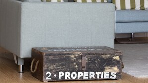 Ein Wohnzimmer mit einem hellblauen Sofa, gestreiften Kissen, einem gerahmten Gemälde und einem Vintage-Koffer mit der Aufschrift "2 PROPERTIES" auf dem Boden.