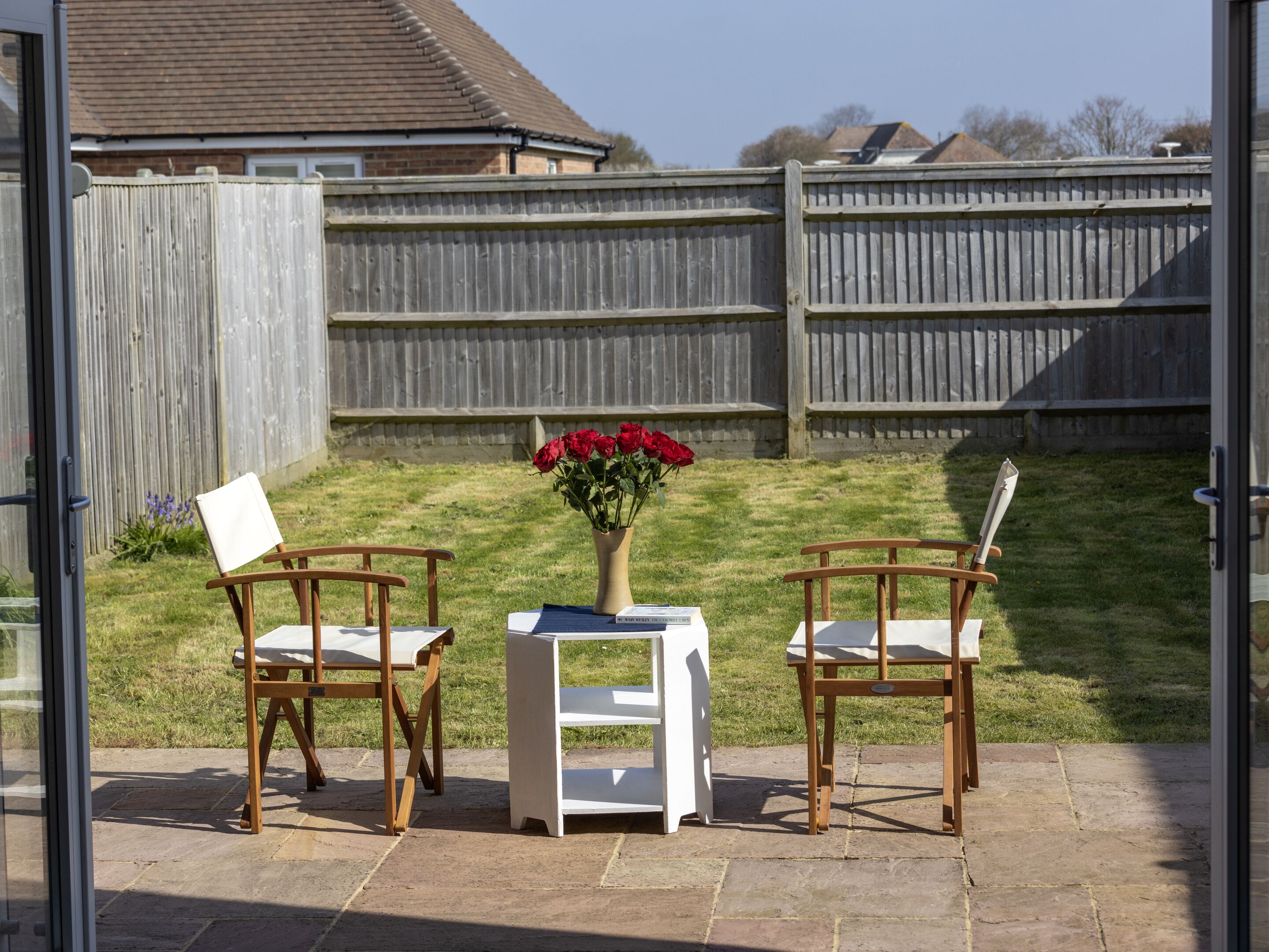 An outdoor patio with two wooden chairs and a small table, a vase with red flowers, and a view of a wooden fence and houses in the background.