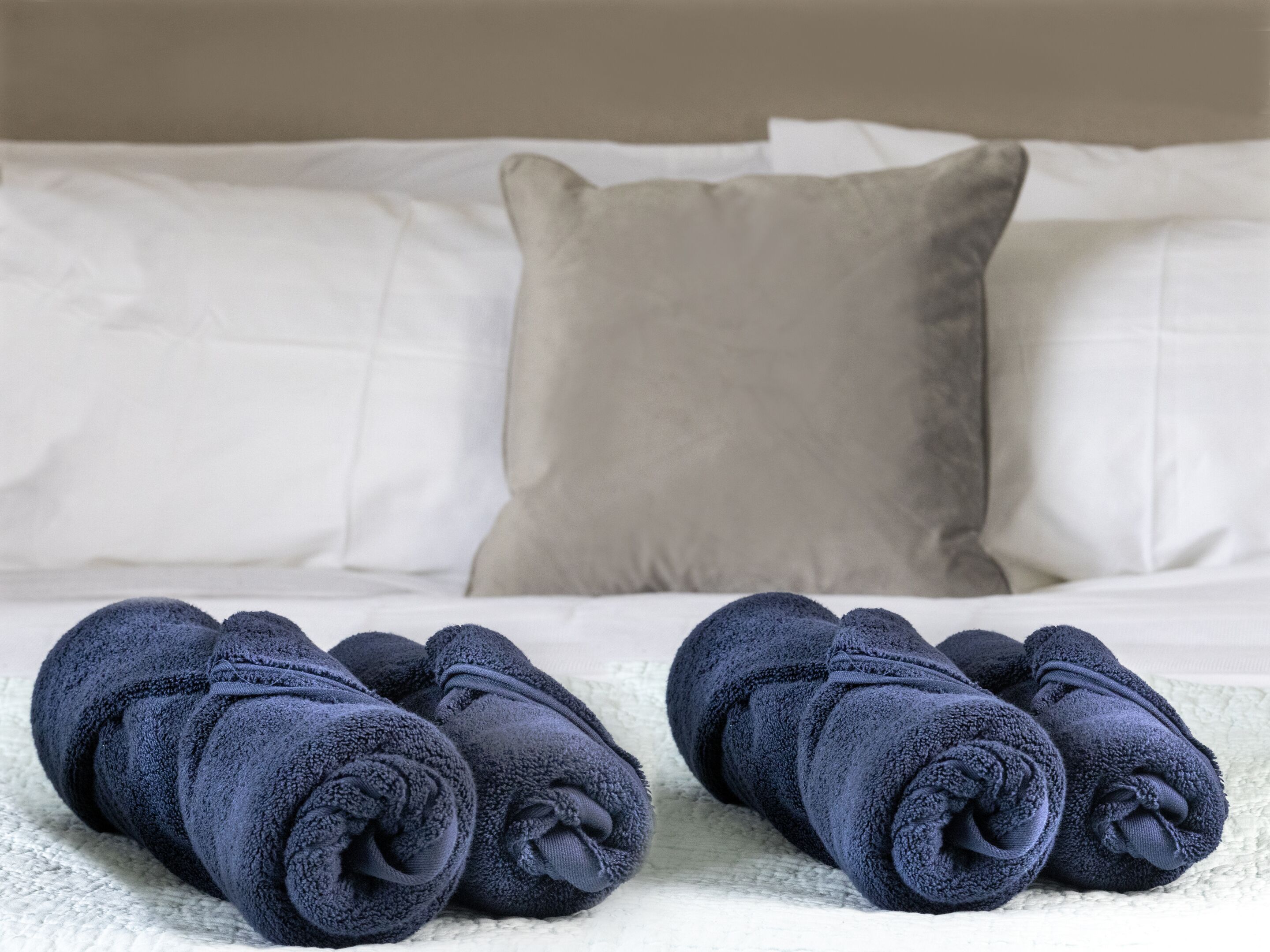 A neatly made bed with white linens and two rolled-up dark blue towels placed on a textured white surface.