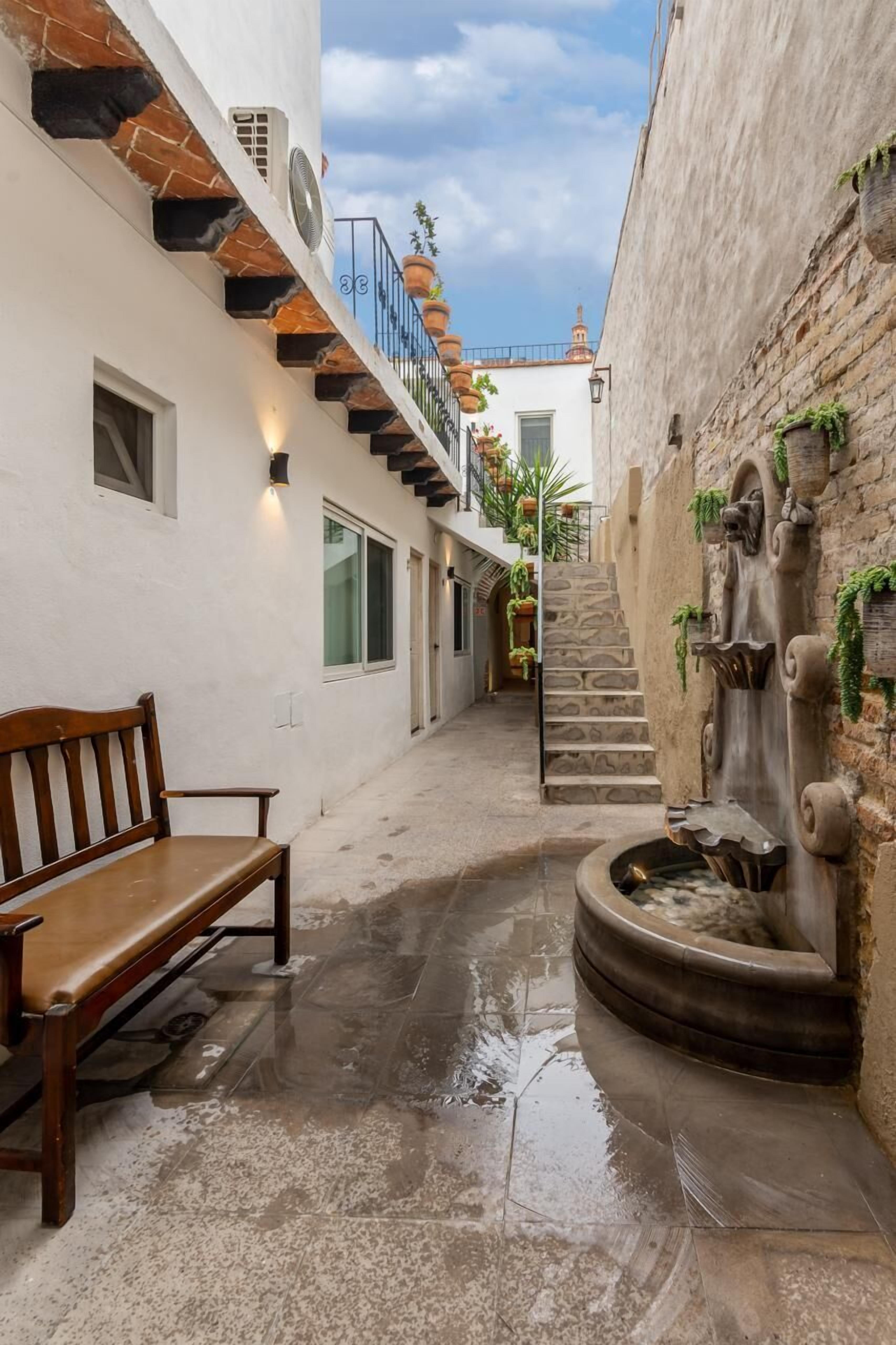 A narrow alleyway with a wooden bench, a stone fountain, and a staircase leading to a second-floor balcony with potted plants.