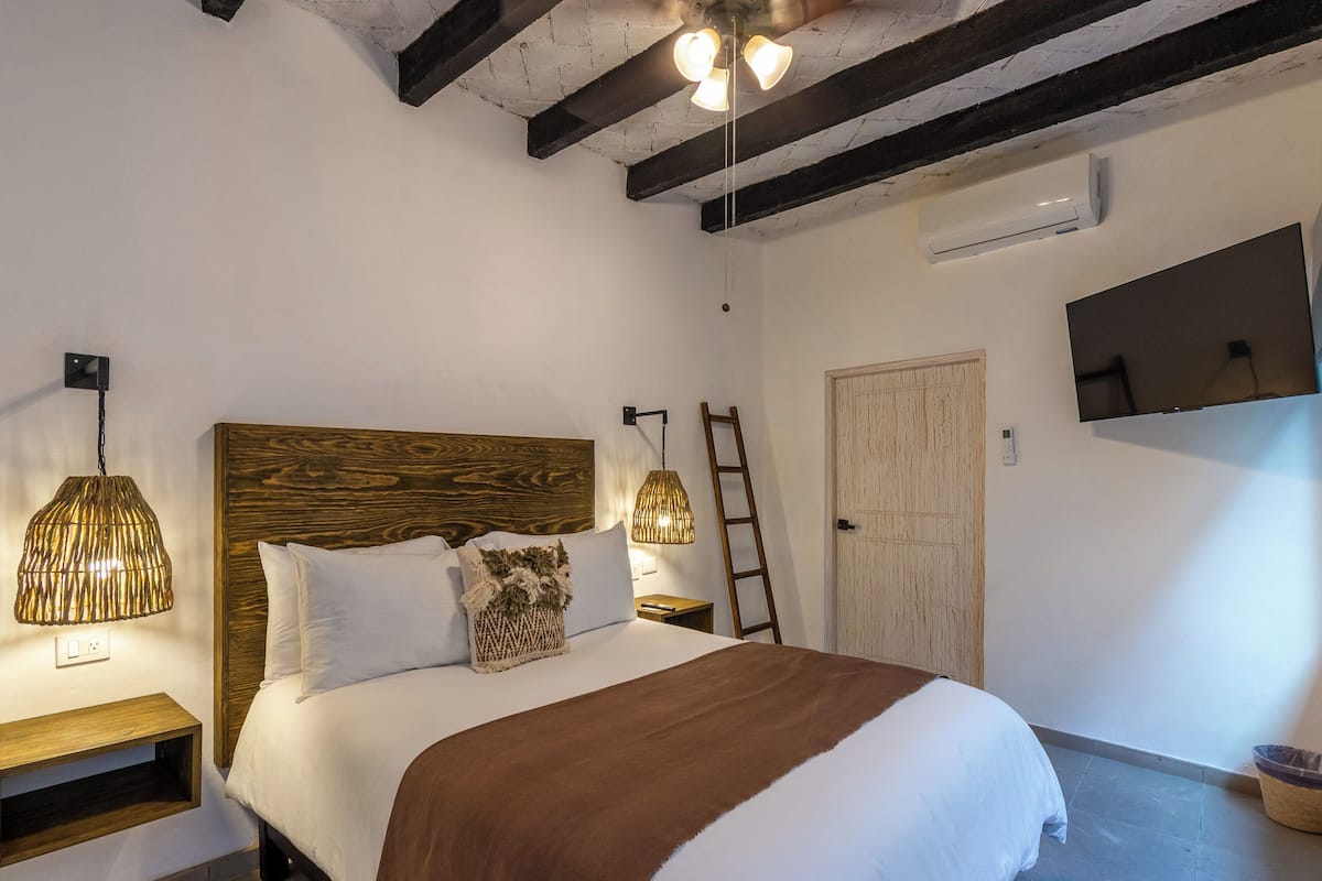 Ein Schlafzimmer mit einem Holz-Kopfteil, einem Bett mit weißer und brauner Bettwäsche, zwei Wandleuchten, einer Leiter und einem Flachbildfernseher.