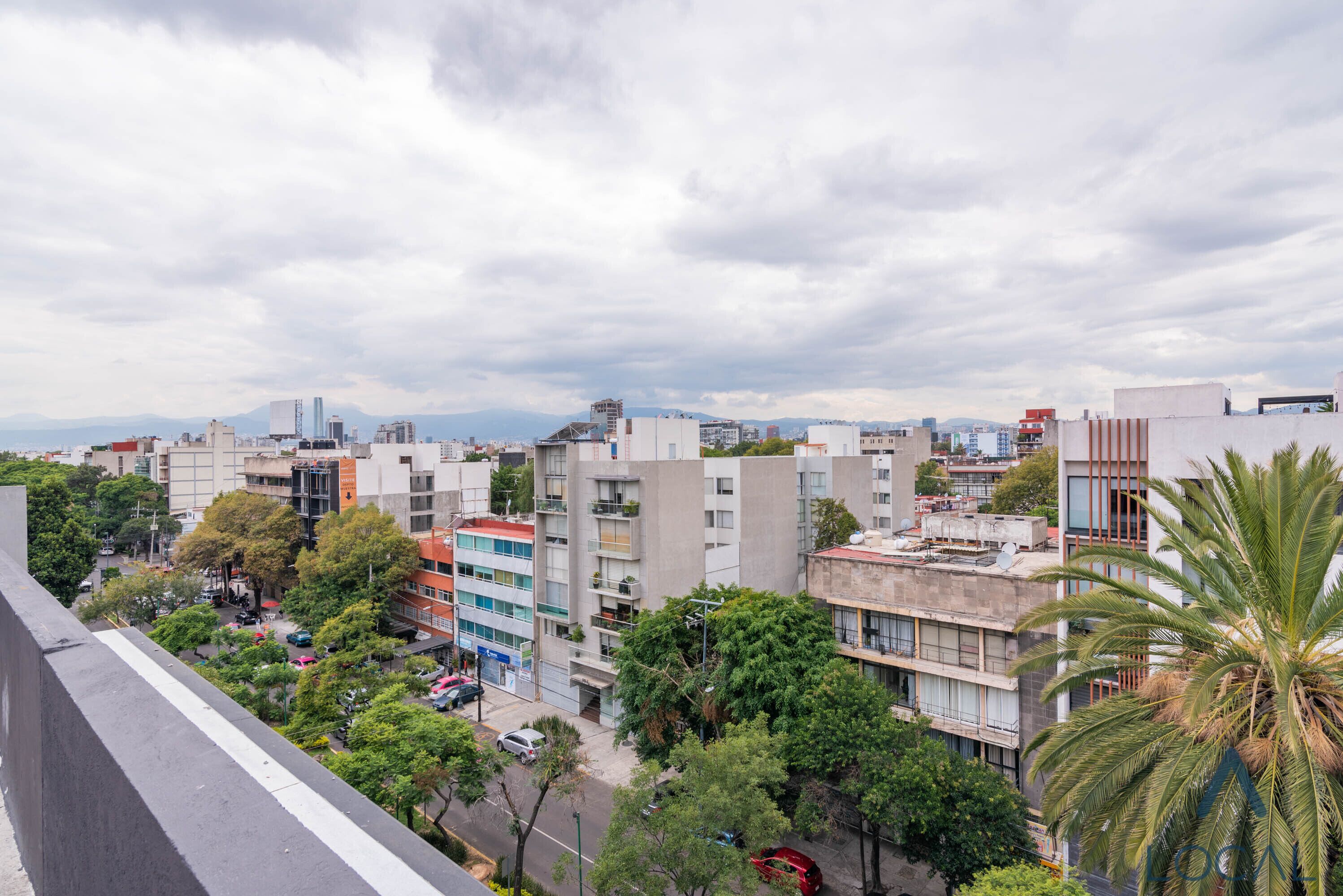 Un paisaje urbano con edificios modernos, una combinación de estructuras residenciales y comerciales, y una vista de montañas distantes bajo un cielo nublado.