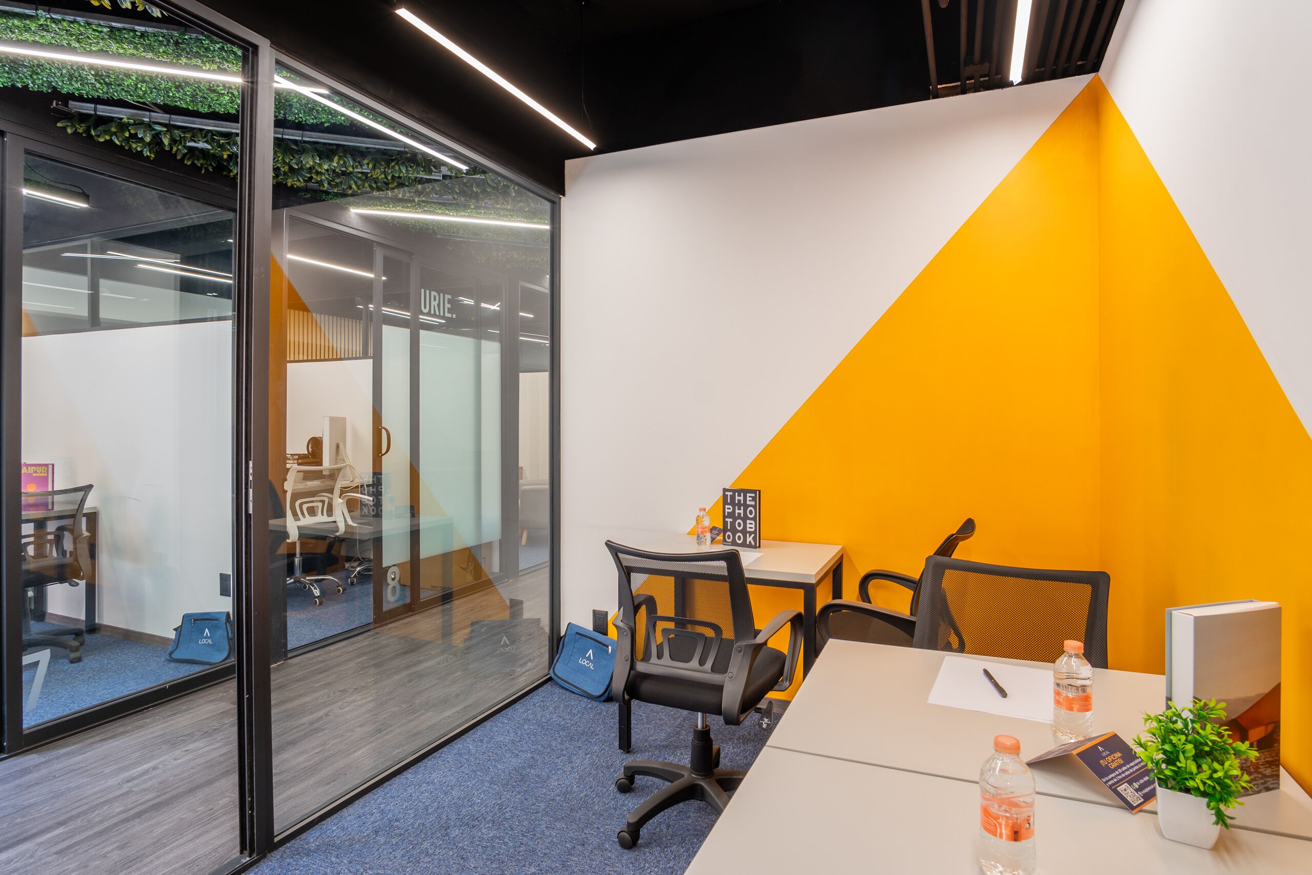 Un espacio de oficina con un separador de vidrio, una pared blanca y naranja, un escritorio con sillas, una computadora y una planta pequeña.