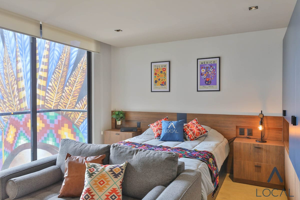 Ein modernes Schlafzimmer mit Bett, Sofa, Nachttisch und Fenster mit farbenfroher Aussicht.