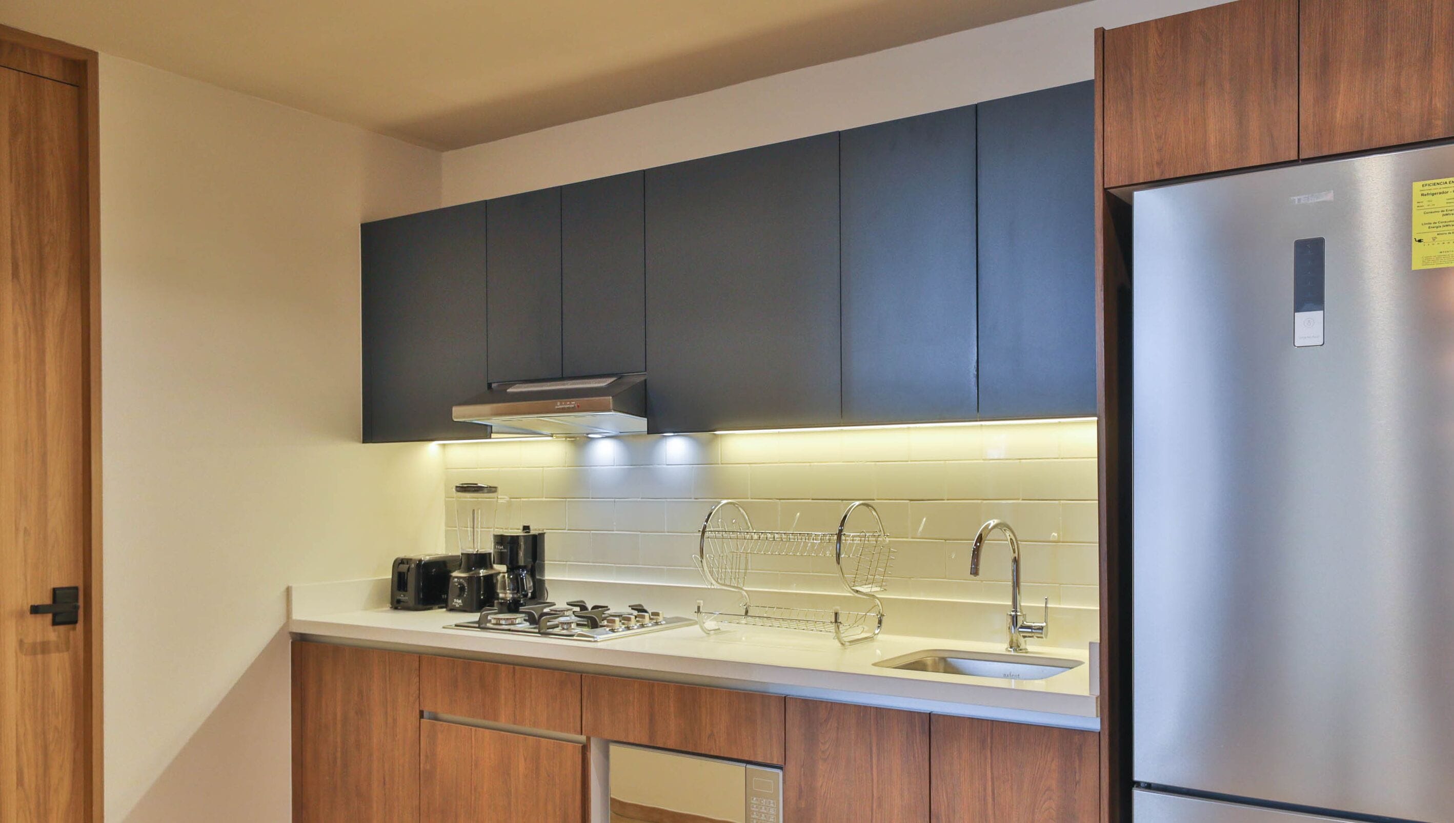 A modern kitchen with wooden cabinets, a stainless steel refrigerator, a built-in oven, a coffee maker, and a sink with a wire rack.