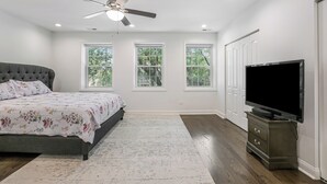 Un dormitorio con una cama, televisión, ventilador de techo y tres ventanas.