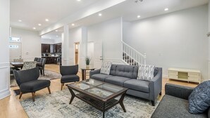 Un salón amplio con un sofá gris, dos sillones, una mesa de centro de vidrio y una escalera que lleva a un piso superior.