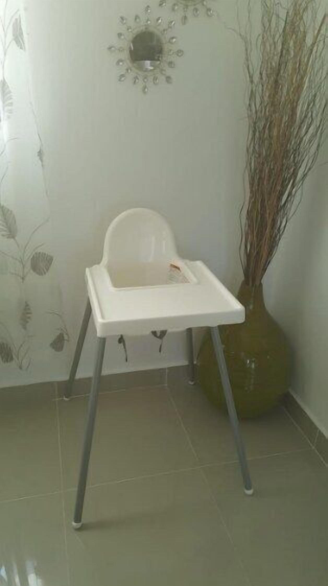 A white baby high chair with a tray table, positioned in a room with a tiled floor and a wall-mounted decorative item.