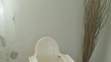 A white baby high chair with a tray table, positioned in a room with a tiled floor and a wall-mounted decorative item.