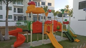 Un parque infantil con toboganes y estructuras para escalar, rodeado de una valla y palmeras.