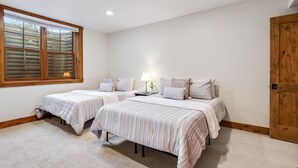 Une chambre avec deux lits, une fenêtre avec des stores, une table de chevet avec une lampe et une porte en bois.