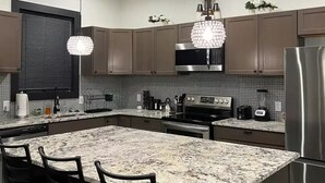 Une cuisine moderne dotée d’un grand îlot central, de meubles en bois foncé, d’appareils électroménagers en acier inoxydable et d’un lustre original.