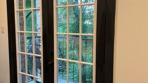 Une porte à cadre noir avec des panneaux de verre blancs, donnant sur un balcon agrémenté d’un mur en pierre et de verdure.