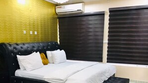A bedroom with a black tufted headboard bed, white bedding, a black coffee table, and a black leather chair. - T Golden Villa Hotel (Enugu)