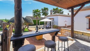 Une terrasse extérieure avec une table en bois et des tabourets de bar, un toit de chaume et une vue sur un bâtiment blanc doté d’un toit en tuiles.