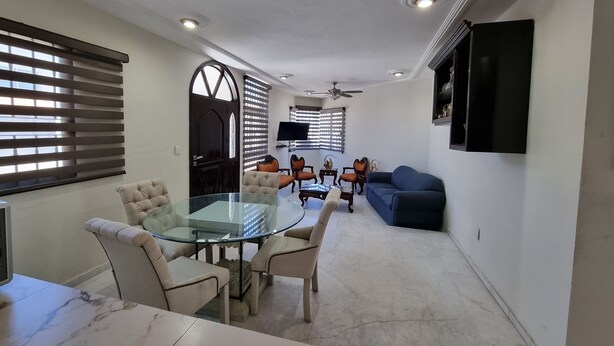 A modern living room with a glass dining table, beige upholstered chairs, a blue sofa, and a flat-screen TV mounted on the wall.