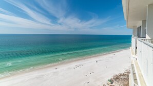 A beach view from a balcony overlooking a sandy beach with clear blue water and a few people.