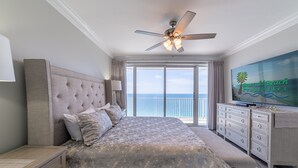A bedroom with a large bed, a ceiling fan, a TV, and a view of the ocean.