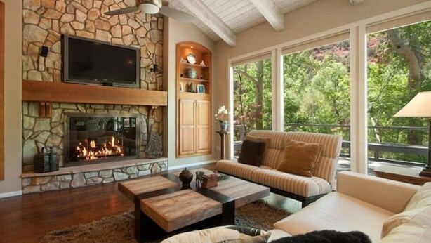 Una sala de estar acogedora con chimenea de piedra, un televisor de pantalla plana y un amplio ventanal con vistas a un jardín exuberante.