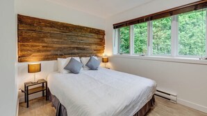 Un dormitorio con cabecera de madera, una cama grande con ropa de cama blanca, dos lámparas de noche y una ventana con vistas a árboles.