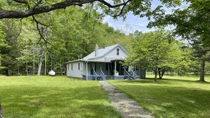 Une petite maison à un seul étage, dotée d’un porche et d’un chemin menant à l’entrée, entourée d’une végétation luxuriante et d’arbres.