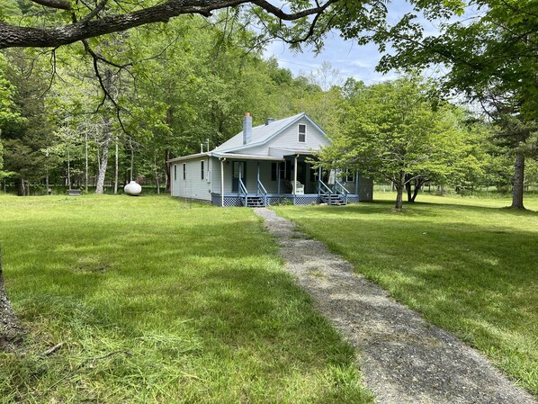 A small, single-story house with a porch and a pathway leading to it, surrounded by lush greenery and trees.