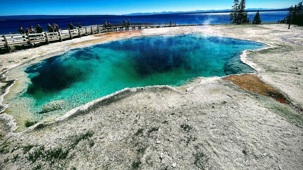 Vista despejada de un manantial termal geotérmico con una plataforma de madera y una cadena montañosa distante.