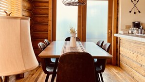 Interior de una cabaña de madera con una mesa de comedor, sillas, una lámpara y vistas a través de las ventanas.