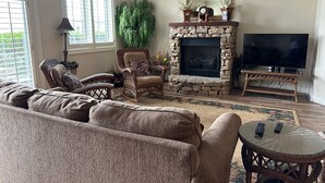A cozy living room with a stone fireplace, a large flat-screen TV, wicker furniture, and a large green plant. - Lakeside Condo ~ Close to Everything ~ First Floor ~ Pool ~ Dock ~ Views (Guntersville)